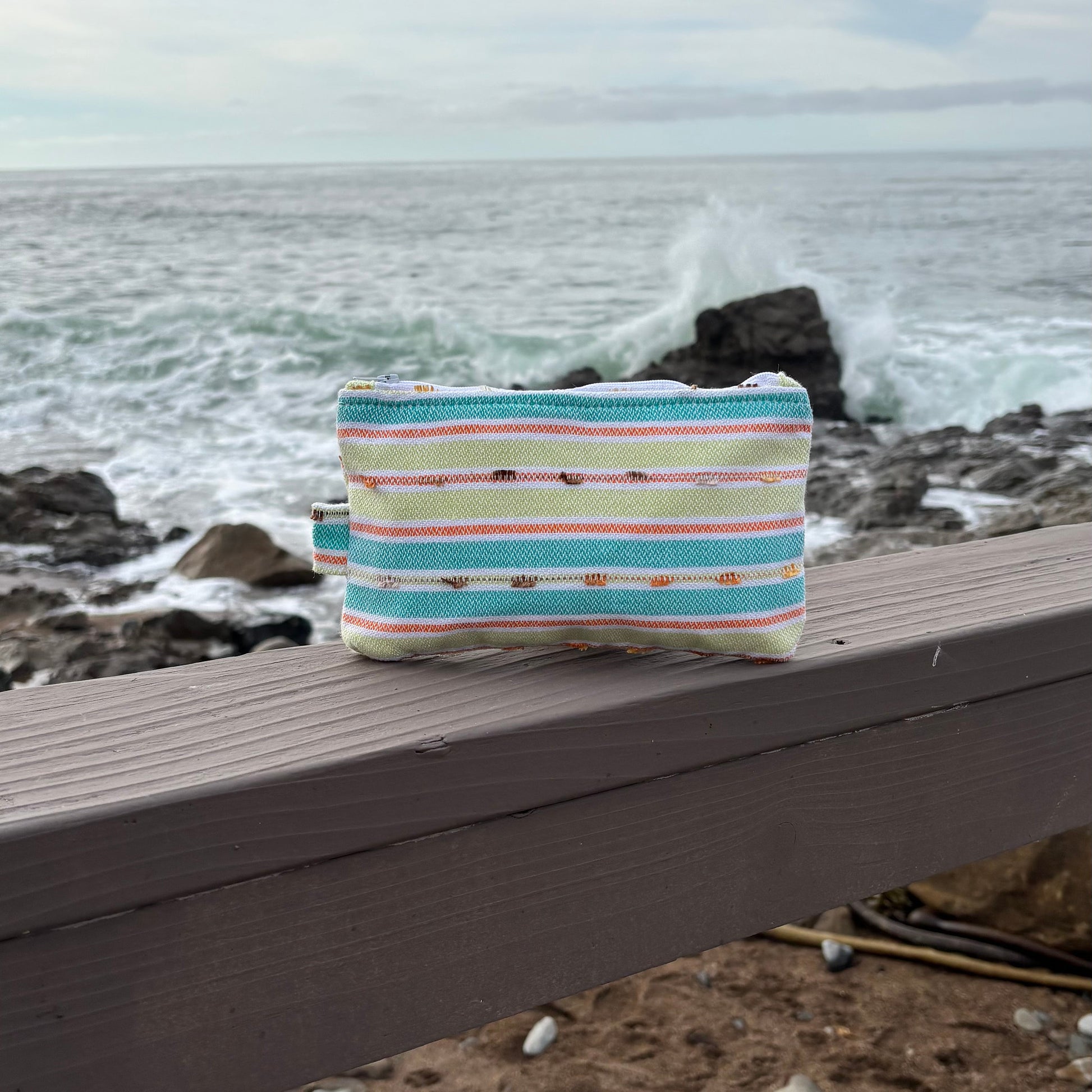Small makeup bag with vibrant  green striped patterns placed on a wooden surface near a coastal backdrop.