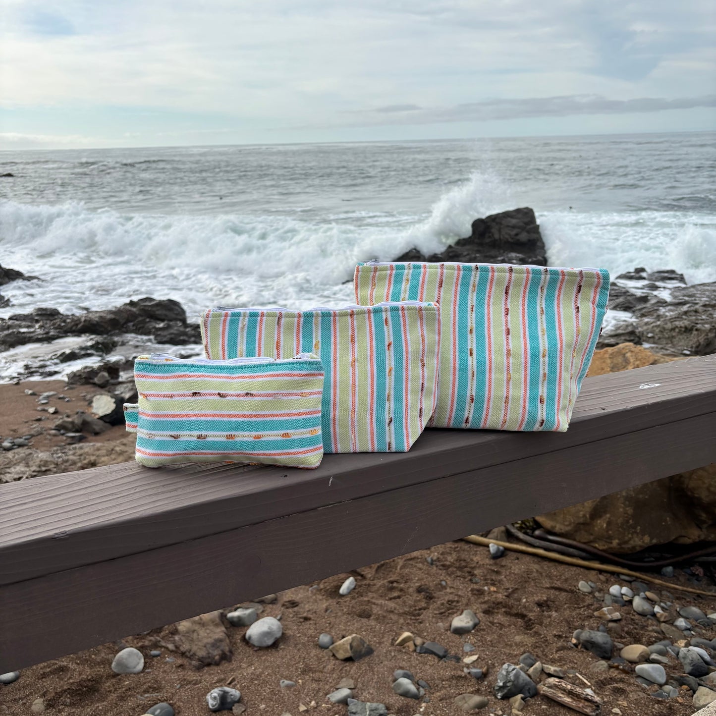 Makeup bags in various sizes with green vibrant striped patterns, placed on a wooden surface near a coastal backdrop.