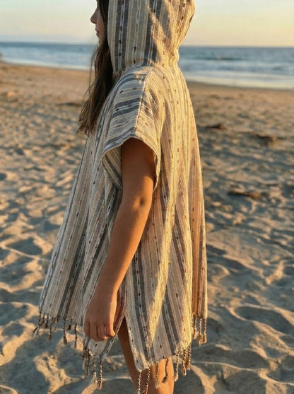 Child wearing a striped, beige and tan hooded poncho with tassels on a beach at sunset.