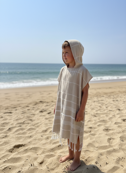 Child wearing a beige hooded poncho on a sandy beach with ocean in the background
