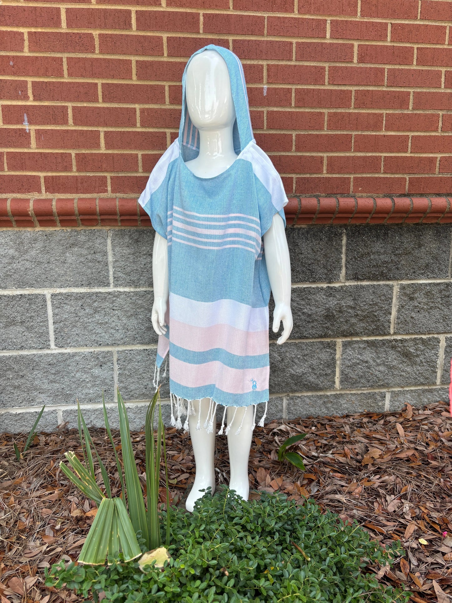 Mannequin wearing a poncho with blue, pink and white stripes on a sidewalk with a brick wall and plants in the background 