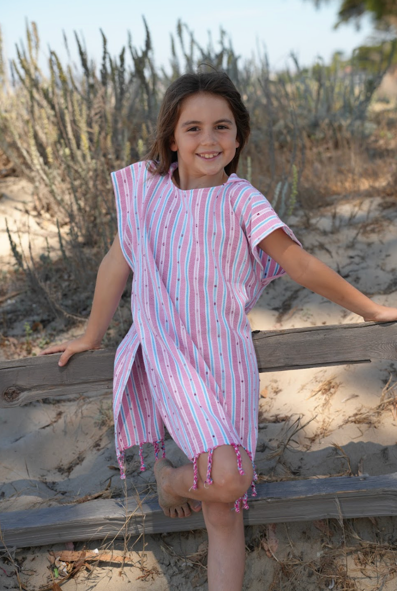 A child wearing a pink striped hooded sun poncho with tassel detailing, standing outdoors.