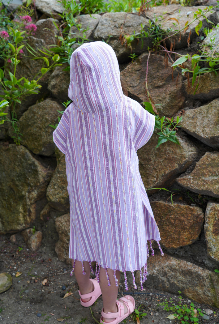 Back view of a child wearing a purple striped hooded sun poncho with tassel detailing, standing outdoors.