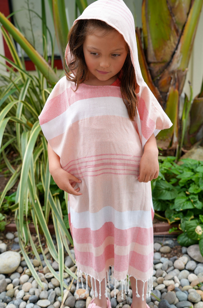A child wearing a peach, red and white striped hooded sun poncho with tassel detailing, standing outdoors.