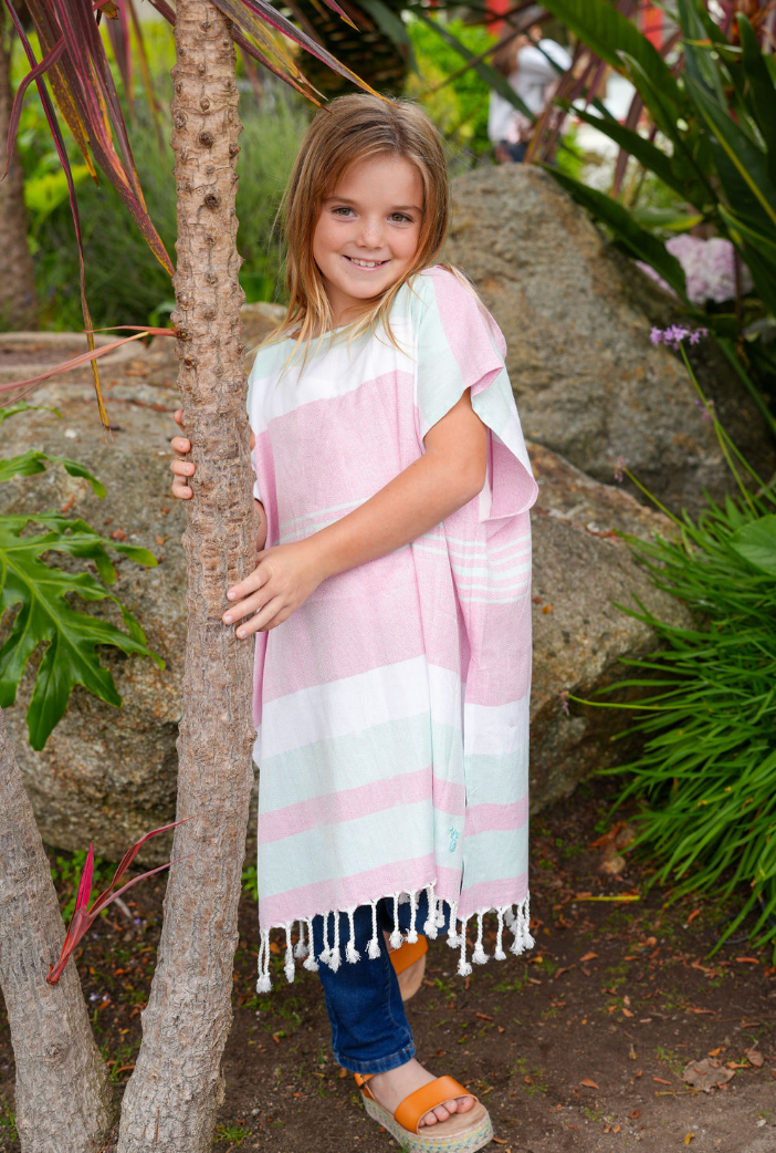 A child wearing a pink, green and white striped hooded sun poncho tassel detailing, standing outdoors.