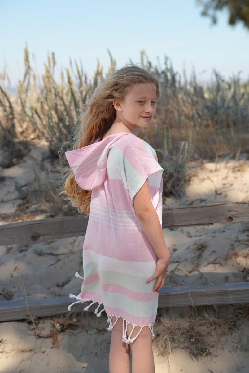 Back view of a Achild wearing a pink, green and white striped hooded sun poncho with tassel detailing, standing outdoors.