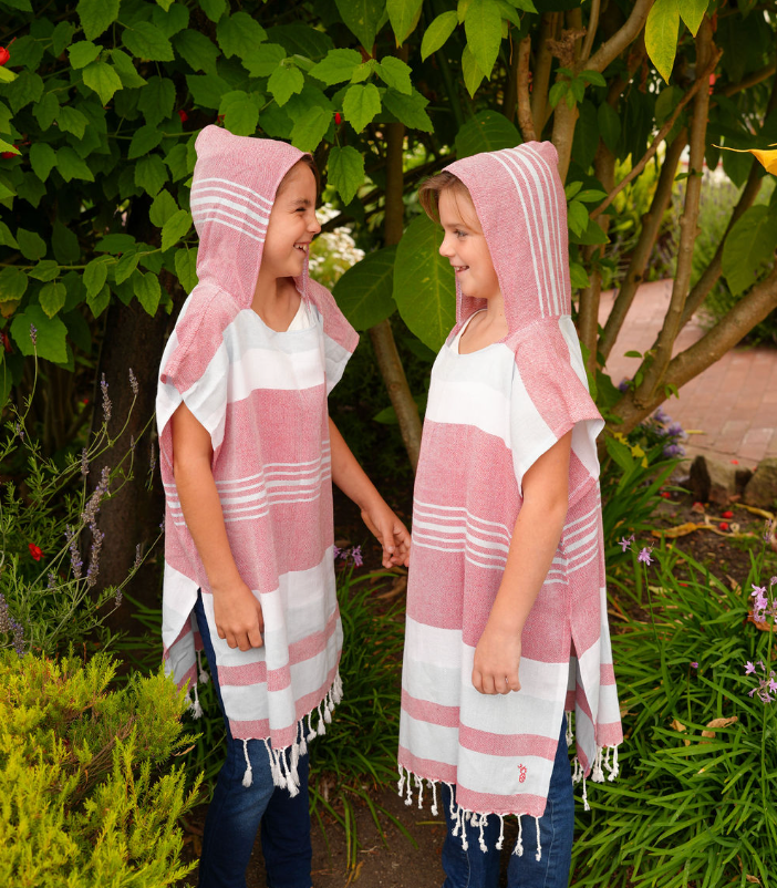 Two children wearing matching red, blue and white striped hooded sun ponchos with tassel detailing, standing outdoors.