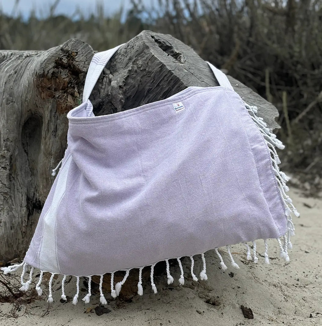 Light purple bag with white fringes draped over a log on a sandy beach.
