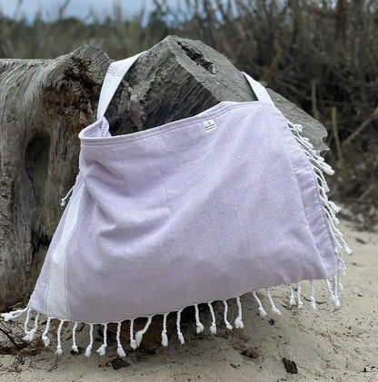 Light purple bag with white fringes draped over a log on a sandy beach.