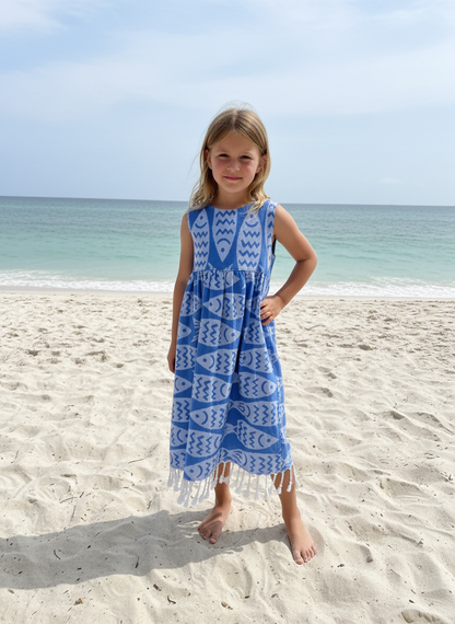 Young girl in a blue dress with white fish patterns standing on a sandy beach.