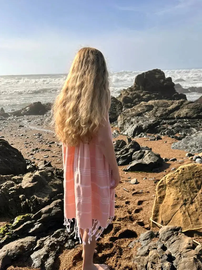 Person wearing a peach dress  standing on a rocky beach with ocean waves.