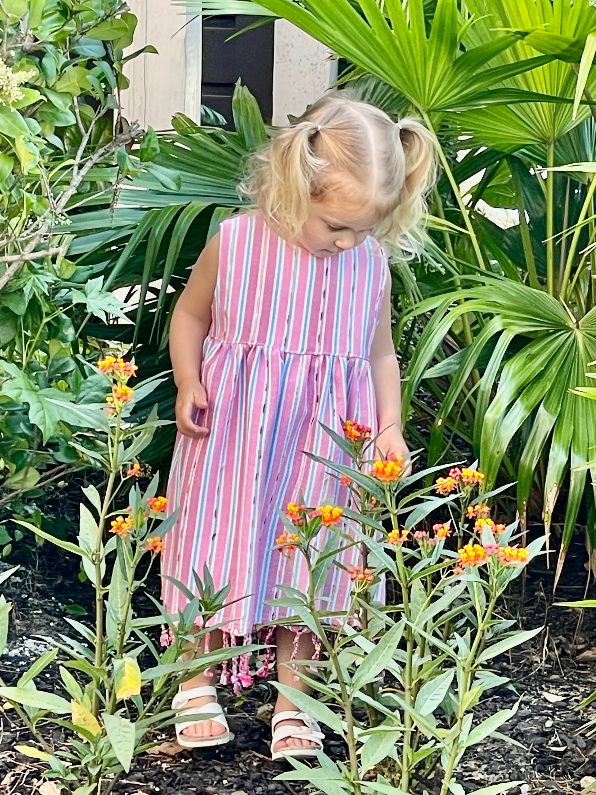 Child in a pink striped dress standing among plants and flowers in a garden setting