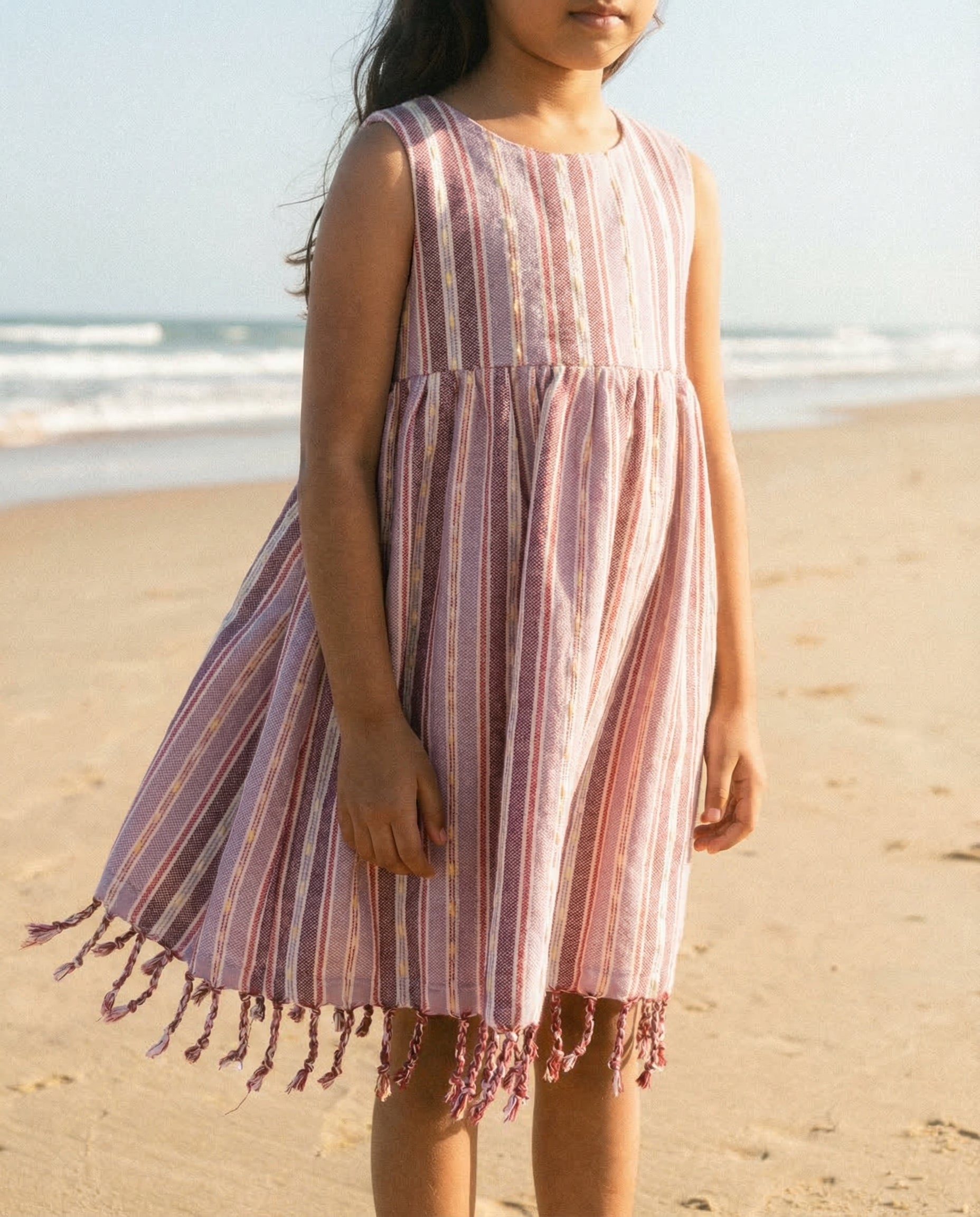 Young girl in a purple striped dress with tassels standing on a beach.