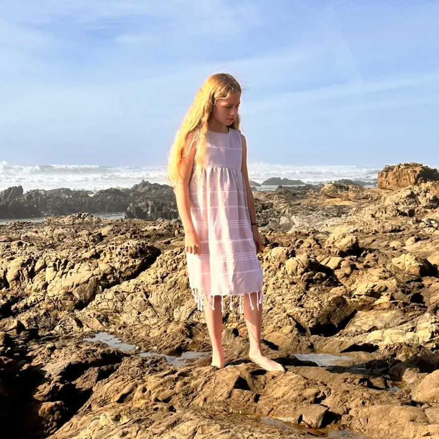 Young girl in a tan striped dress standing on rocky coastal landscape with ocean in the background