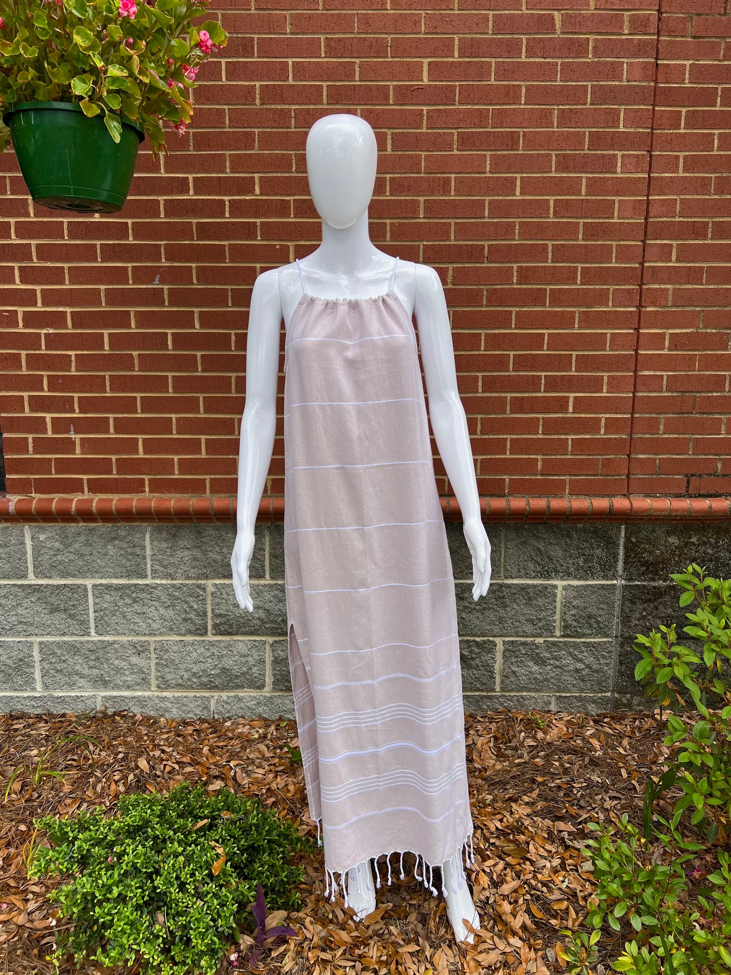 Mannequin wearing tan striped sleeveless maxi sundress with tassels in front of brick wall 