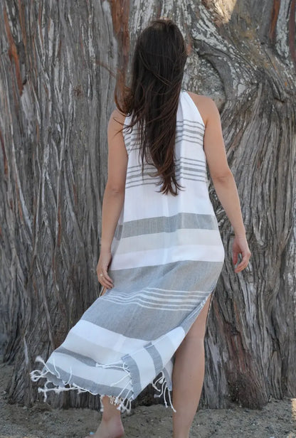 Woman wearing a charcoal, grey and white striped dress with tassels standing against a tree on a beach.