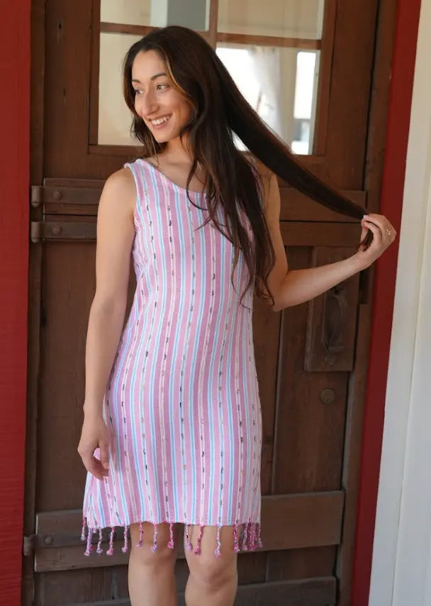 A woman standing outdoors wearing a pink striped sleeveless knee-length dress with  tassels. 