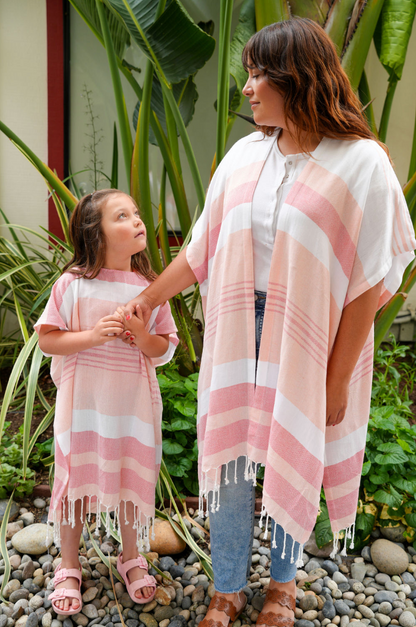 A child wearing a peach, red and white striped hooded sun poncho with tassel detailing, standing outdoors with a woman wearing a matching cover-up over jeans and a white shirt