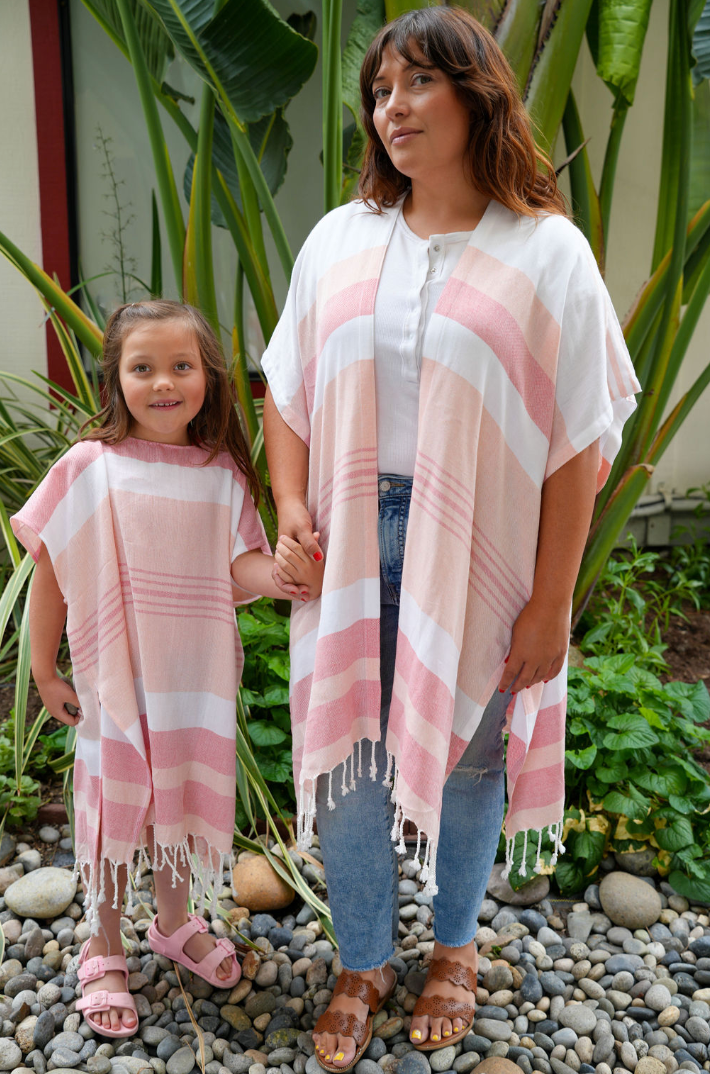 Woman in red, peach and white stripe mid-length cover-up over jeans and a white Tshirt with a girl in a matching poncho in outdoor setting