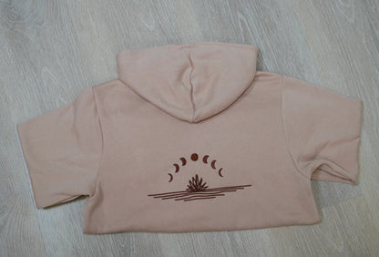 A light brown hoodie laid flat on a wooden surface, with a graphic of the moon in different phases embroidered on the back.