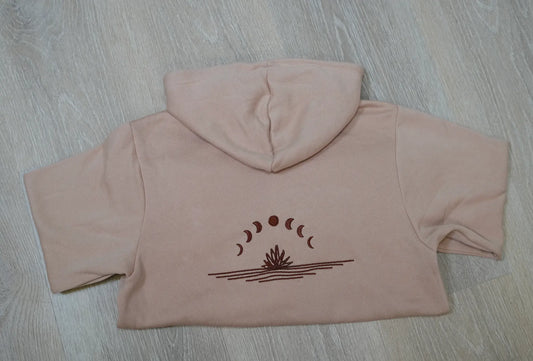 A light brown hoodie laid flat on a wooden surface, with a graphic of the moon in different phases embroidered on the back.