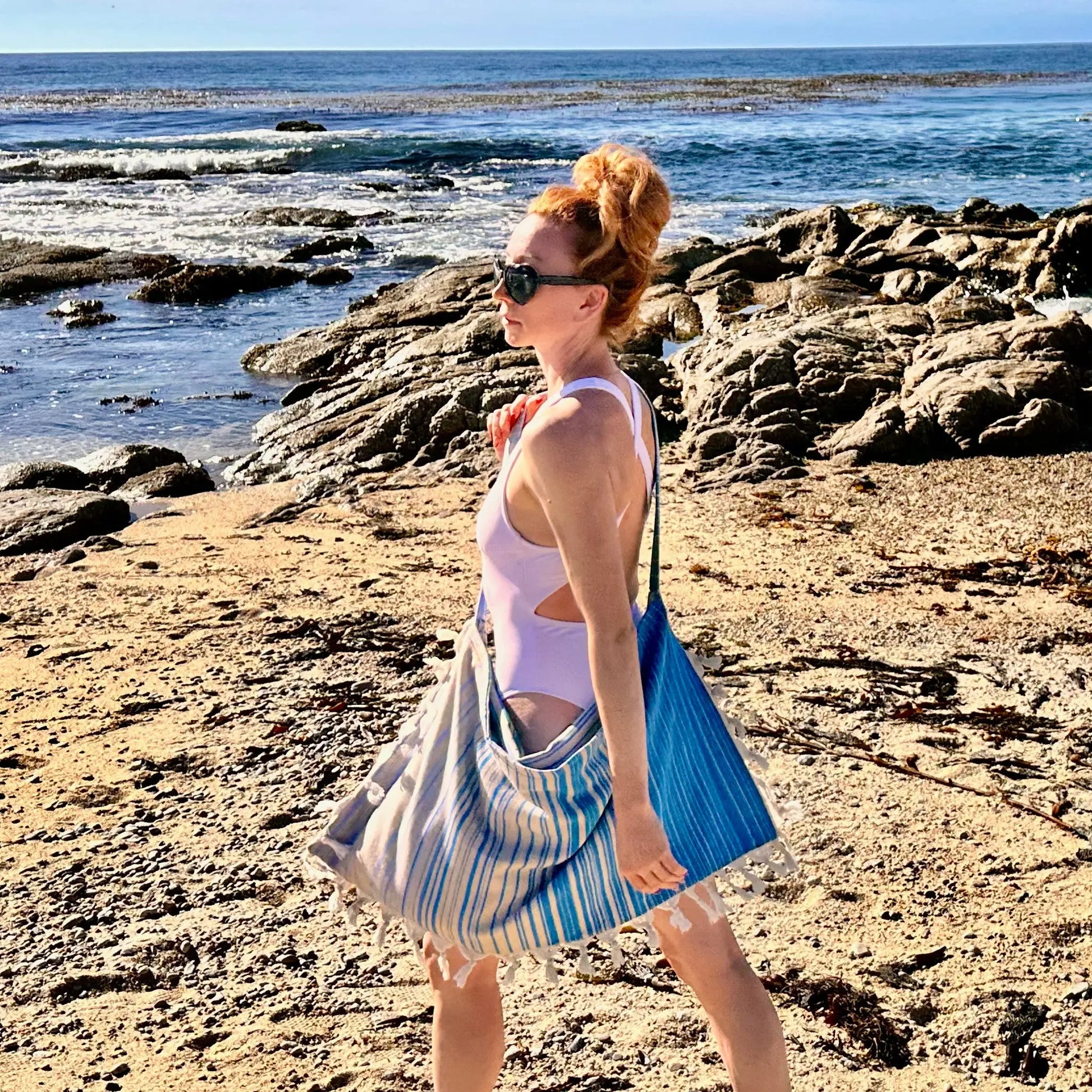 Woman walking on a rocky beach with ocean view carrying a blue striped beach bag