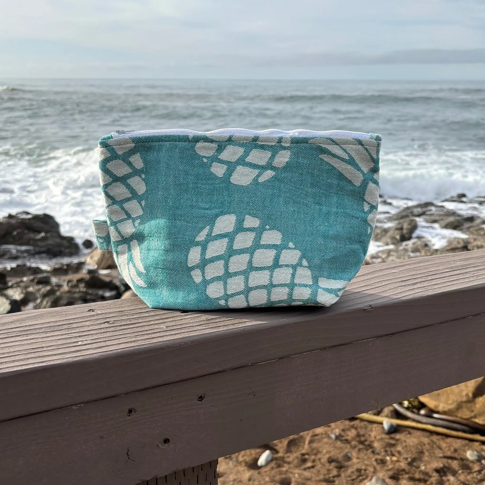 Teal fabric pouch with white pattern on a wooden ledge by the ocean.