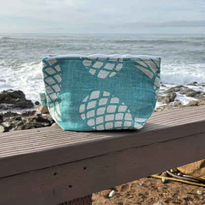 A medium sized  green pineapple print makeup bag placed on a wooden bench with a blurred beach background.