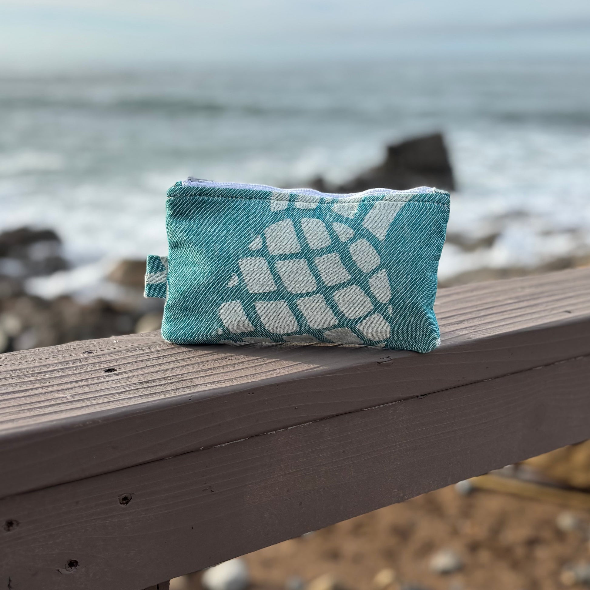 A small green pineapple print makeup bag placed on a wooden bench with a blurred beach background.