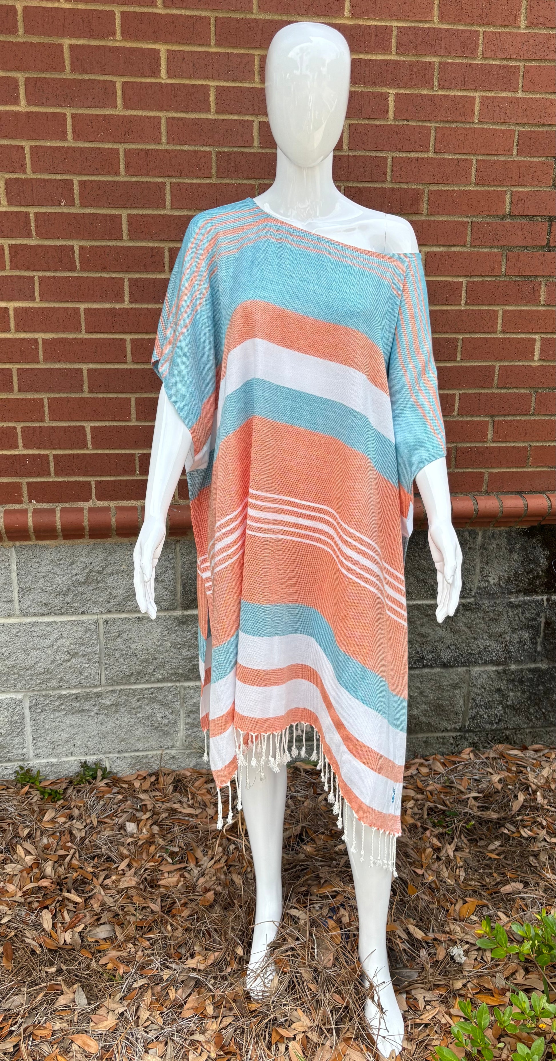 A mannequin displaying a women's poncho in orange, turquoise, and white stripes, with a brick wall and plants in the background.