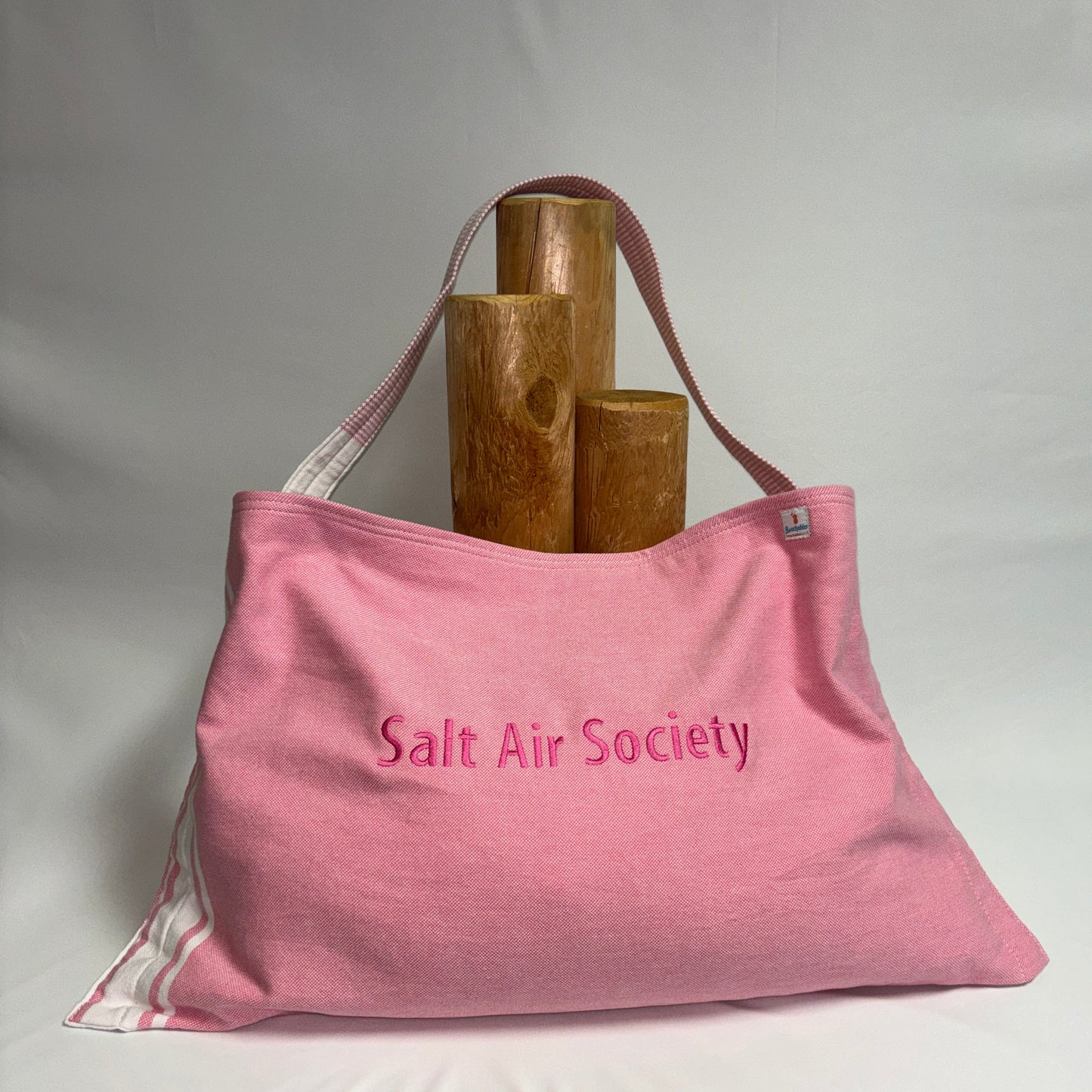 A pink canvas tote bag with 'Salt Air Society' embroidered on the front, displayed on a white draped surface with three wooden cylinder props behind it.