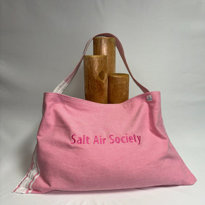 A pink canvas tote bag with 'Salt Air Society' embroidered on the front, displayed on a white draped surface with three wooden cylinder props behind it.