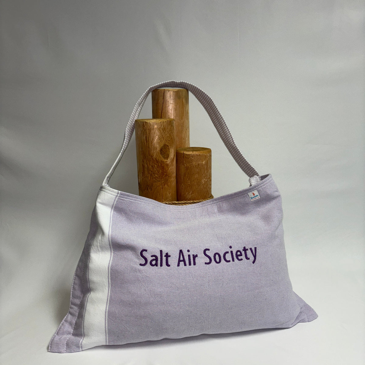 A lavender and white canvas tote bag with 'Salt Air Society' embroidered on the front, holding three wooden candlesticks against a draped white fabric backdrop.