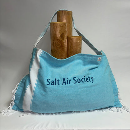 A light blue and white fringed tote bag with 'Salt Air Society' printed on the front, displayed on a white draped surface with three wooden logs behind it.