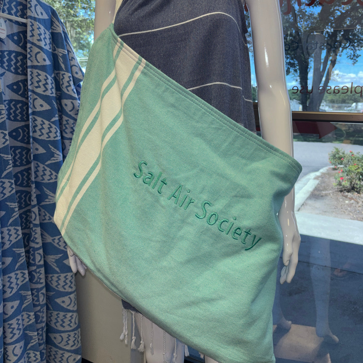 A mint green cotton tote bag with white stripes and the text 'Salt Air Society' embroidered on it, displayed on a mannequin in a store window.