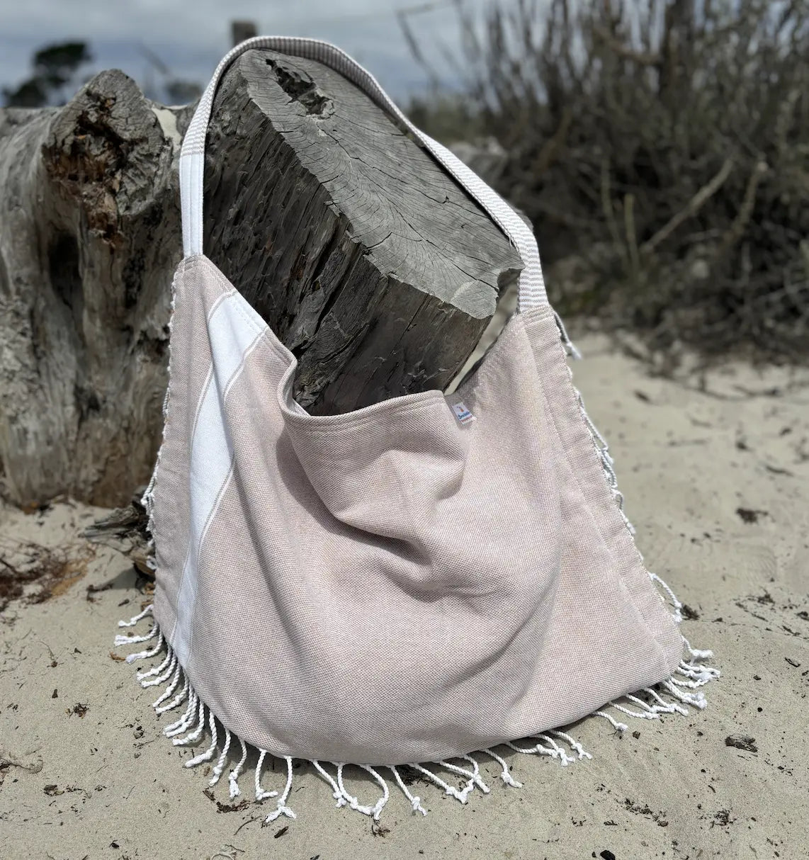 Beige tote bag with white fringe on a sandy beach with driftwood.