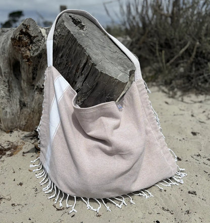 Beige tote bag with white fringe on a sandy beach with driftwood.