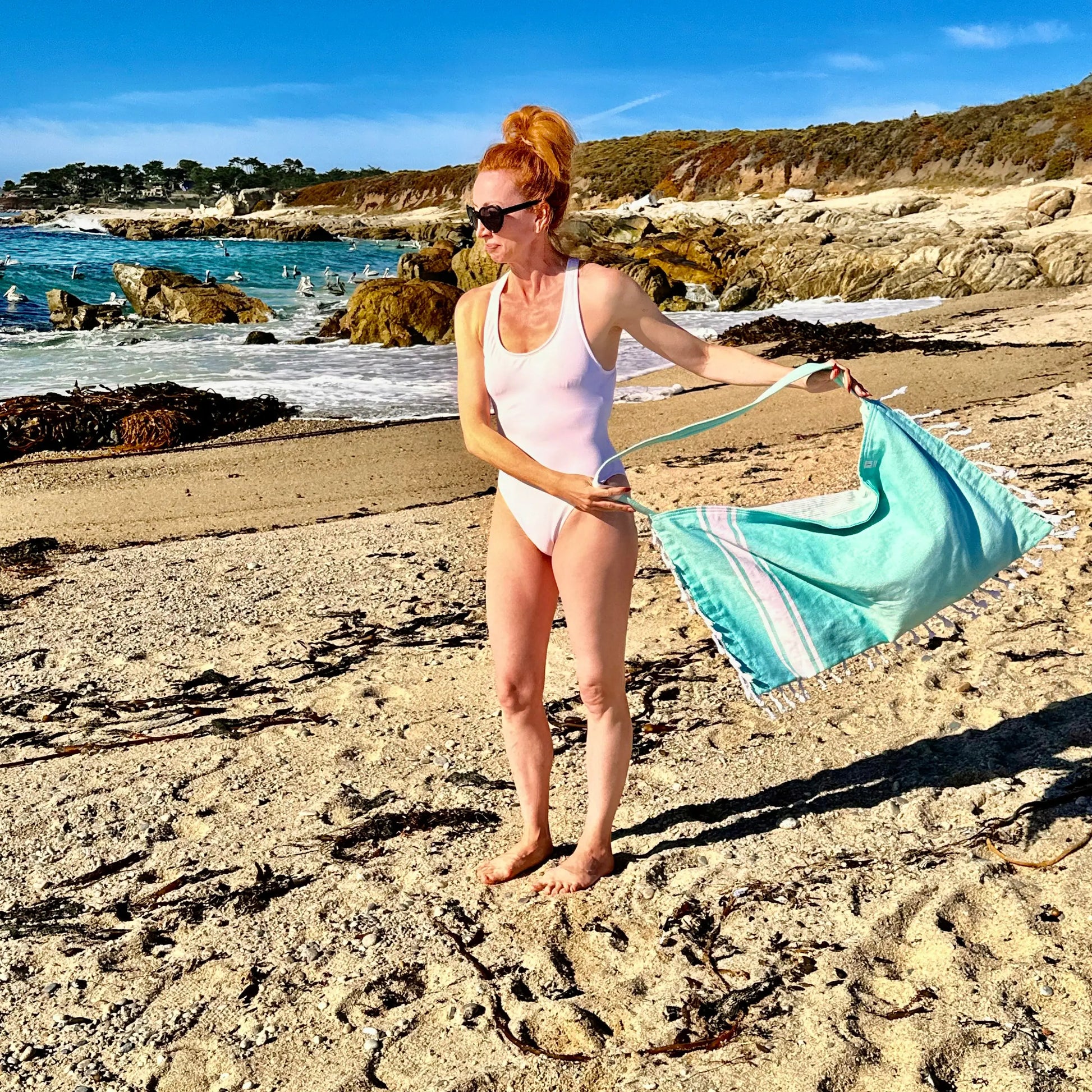 Woman in a swimsuit holding a green bag on a beach with ocean and cliffs in the background