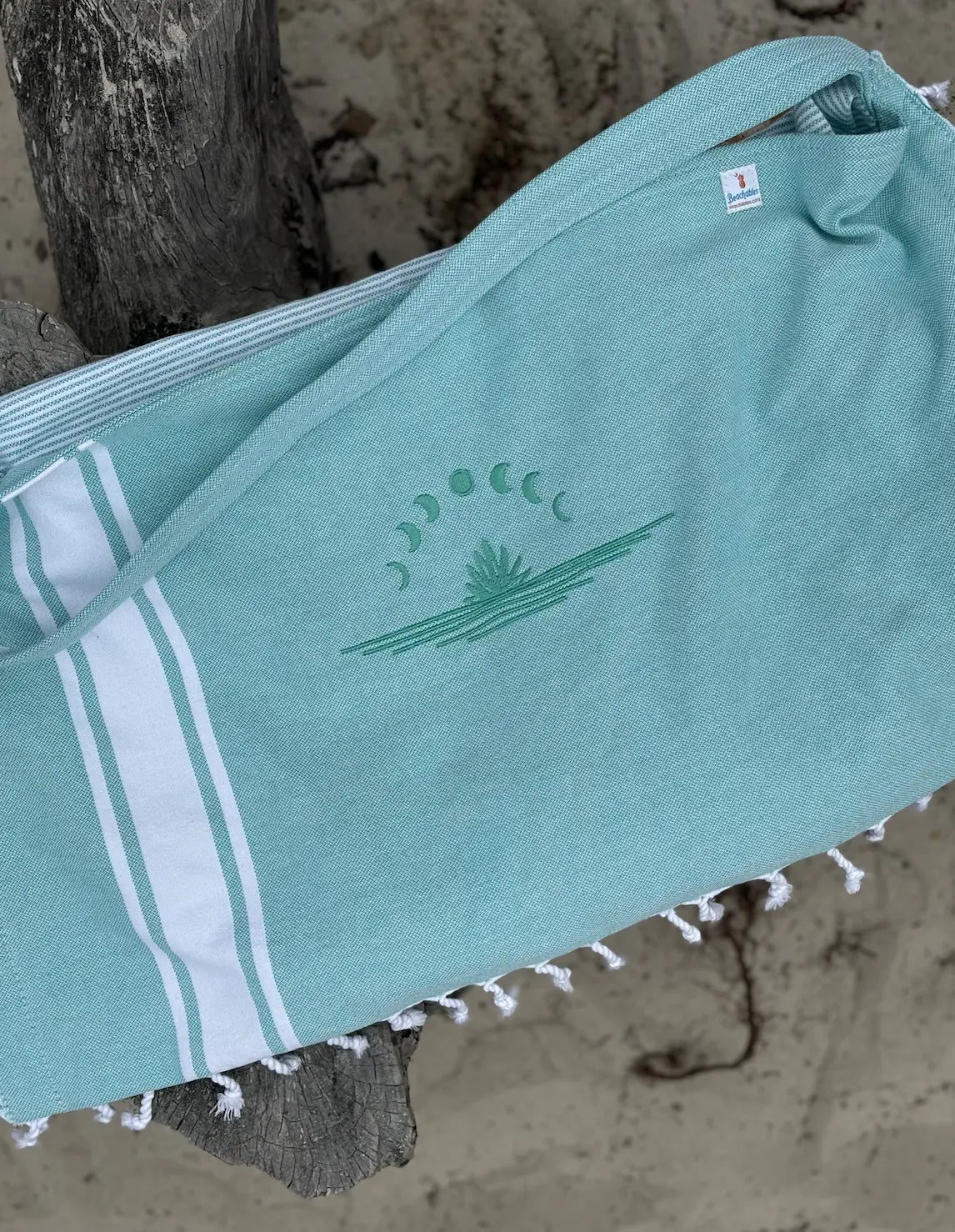 A aqua green and white striped tote bag with a crescent moon and a plant embroidered on the front, lying on a wooden surface.