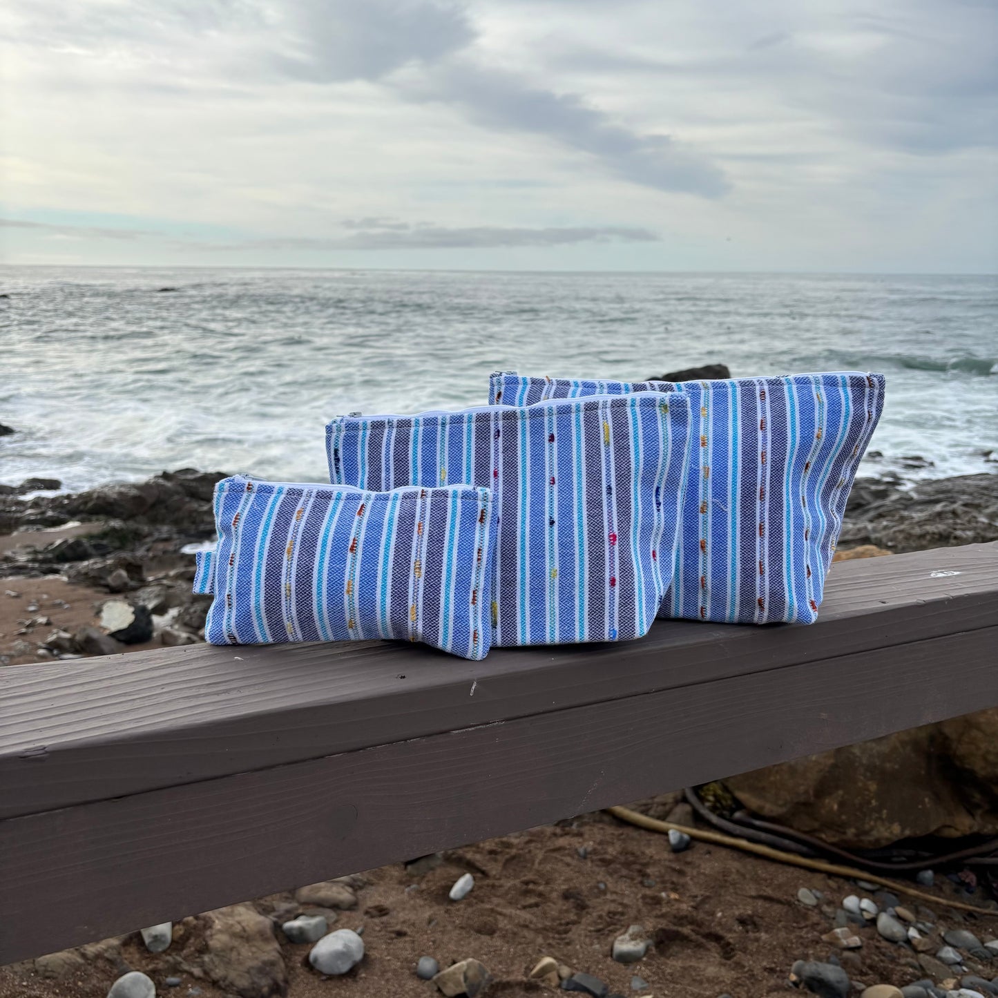 Three blue striped makeup bags in varying sizes placed on a surface with a coastal background.