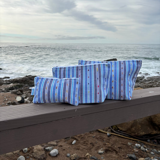 Three blue striped makeup bags in varying sizes placed on a surface with a coastal background.
