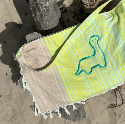 A green and yellow striped tote bag with a dinosaur embroidery on the front, lying on a sandy surface.