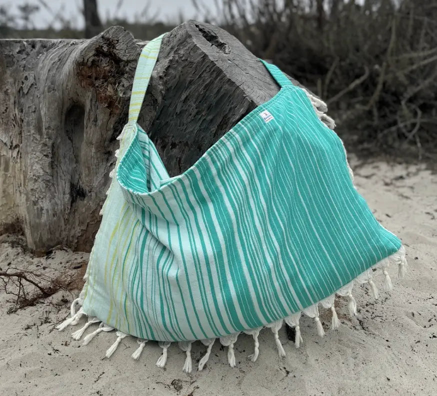 Teal and pale blue striped bag with tassels on a sandy beach.