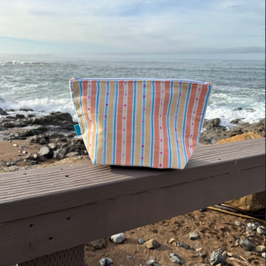 A large sized orange and yellow striped makeup bag placed on a wooden surface with a beach background.