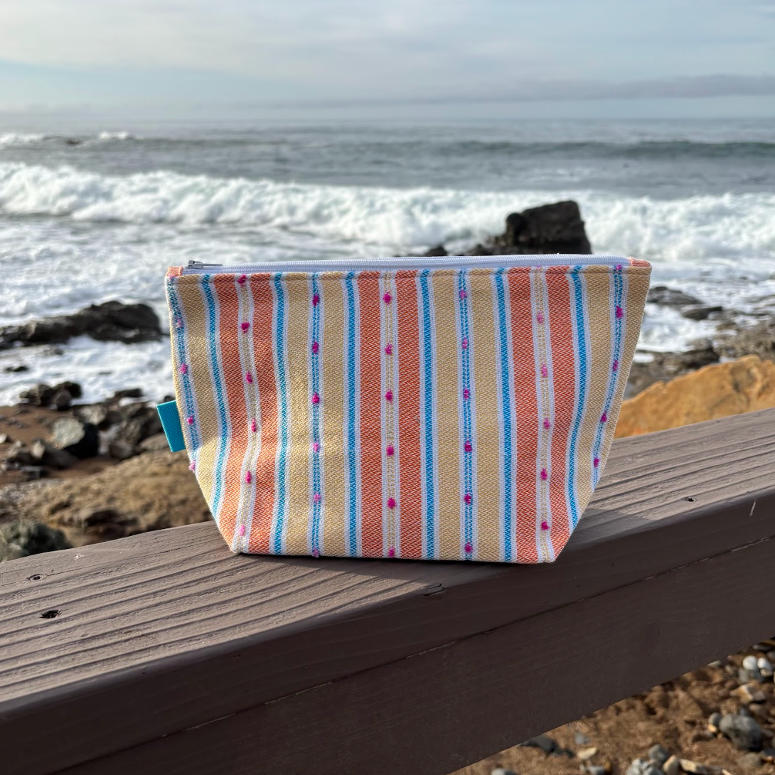 A medium sized orange and yellow striped makeup bag placed on a wooden surface with a beach background.