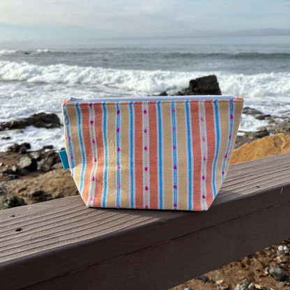 A medium sized orange and yellow striped makeup bag placed on a wooden surface with a beach background.