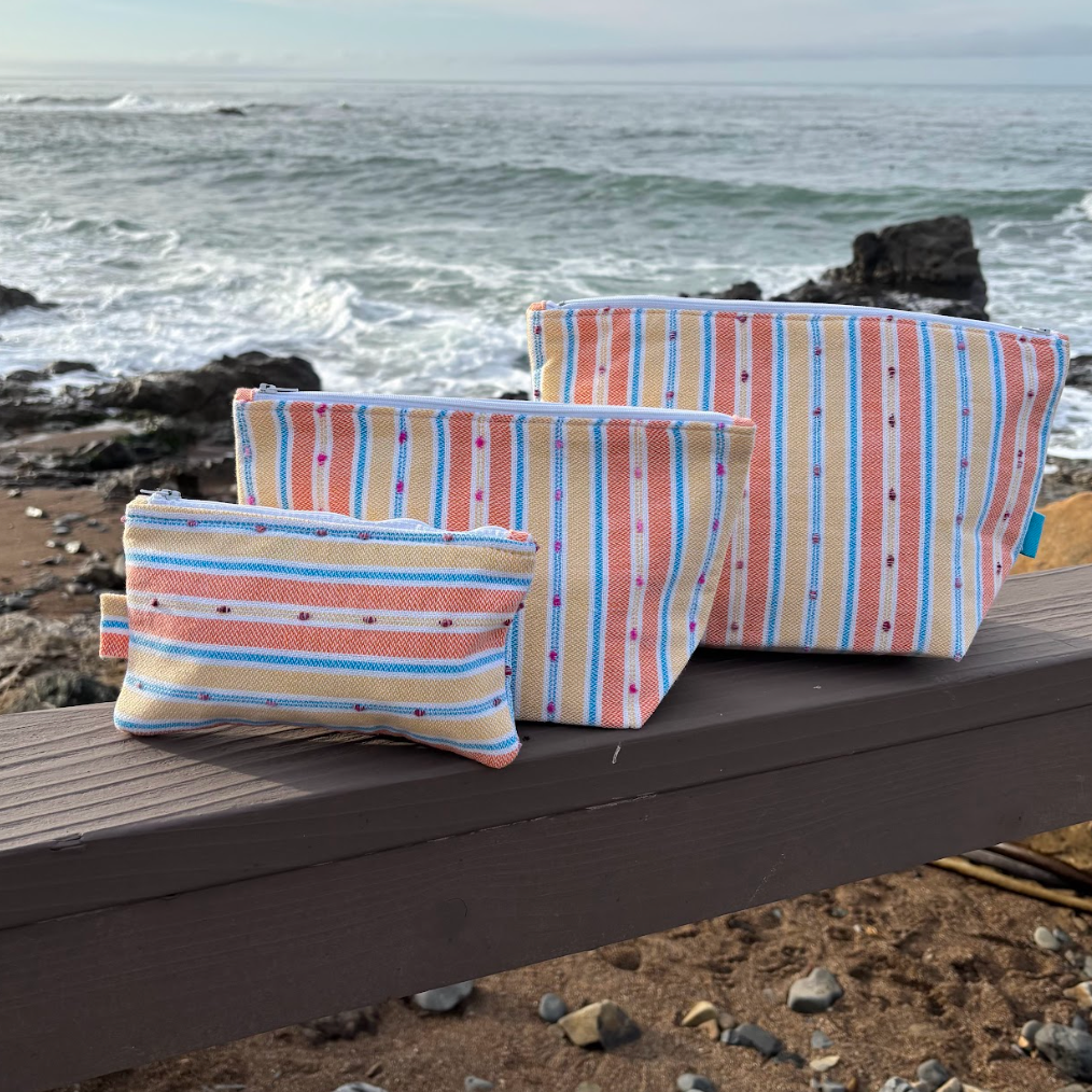 A set of three orange and yellow striped makeup bags in varying sizes placed on a wooden surface with a beach background.