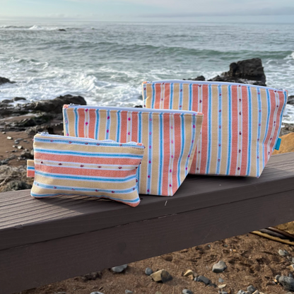 A set of three orange and yellow striped makeup bags in varying sizes placed on a wooden surface with a beach background.