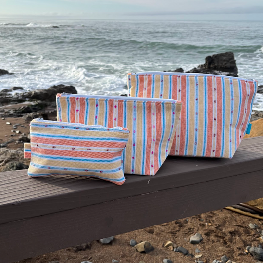 A set of three orange and yellow striped makeup bags in varying sizes placed on a wooden surface with a beach background.