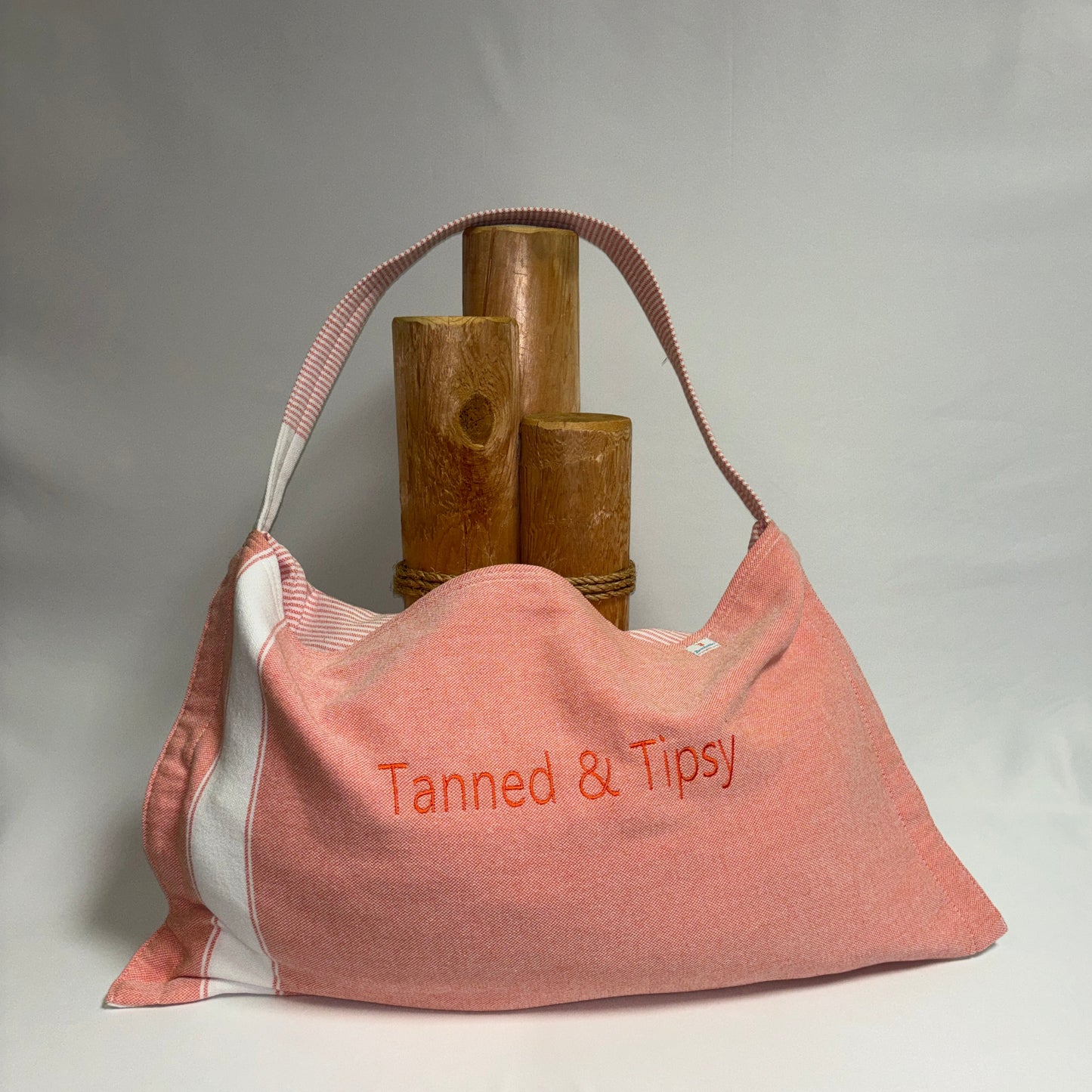 A dusty pink canvas shoulder bag with white side panels and a pink-and-white striped strap, featuring the text 'Tanned & Tipsy' embroidered in orange on the front. The bag is styled with three rustic wooden cylinder props against a draped white fabric backdrop.